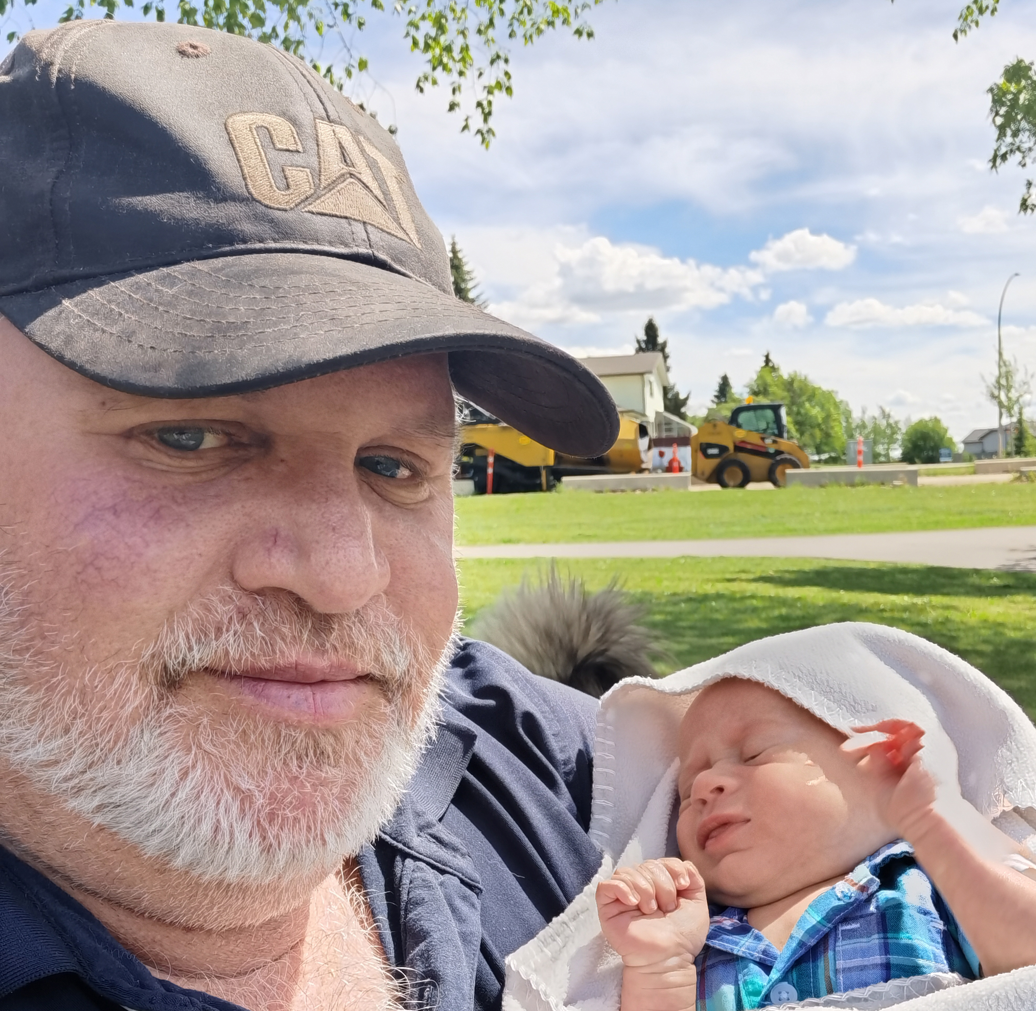 head shot of Roy DG Cooper Protopian Grandmaster. The image in the section “ask a Protopian” is of me, the founder and protopian grandmaster, with a leather CAT baseball cap holding my grandson at about three months old. In the background is a park with a CAT skid steer and CAT paver in the distance.  